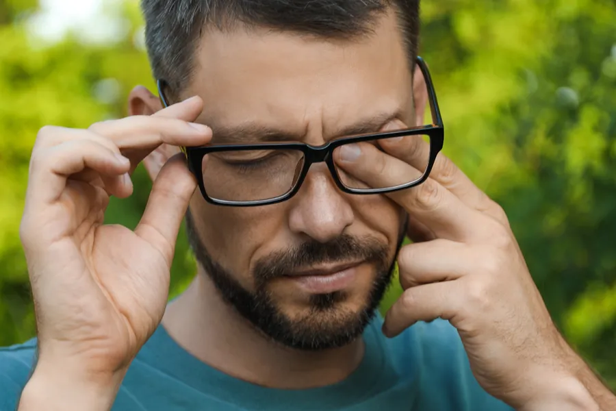 Allergic-Conjunctivitis-Clinic A man with a beard and glasses standing outside and wearing a blue t-shirt lifts his glasses and rubs his eyes due to allergy irritation. Get treatment for allergic conjunctivitis from AZENA Health & Longevity in Gainesville.