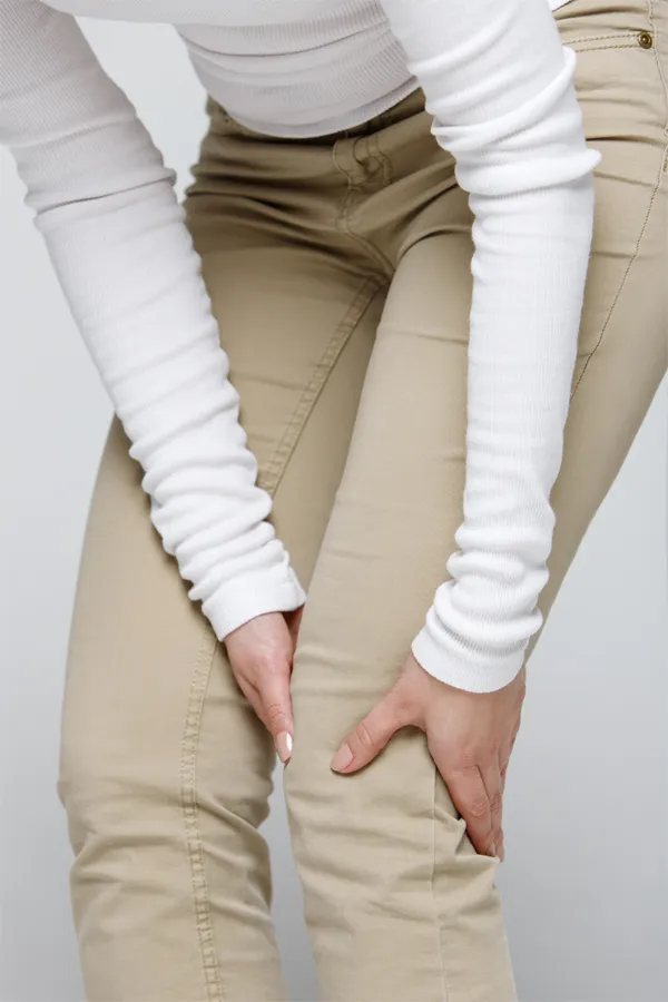 Arthritis-Treatment A woman wearing a white long-sleeve shirt and tan pants, clutching her knee, suffering from arthritis. Get arthritis treatment from AZENA Health & Longevity in Gainesville.