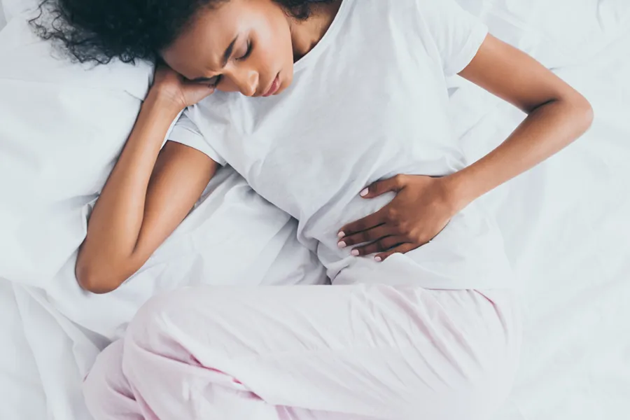 Crohns-Disease-Clinic A dark-skinned woman in white clothes lays on her bed clutching her middle before getting treatment for Crohn's Disease from AZENA Health & Longevity in Gainesville.
