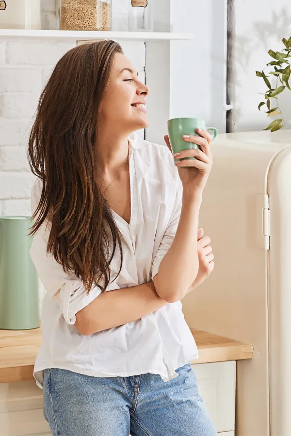 Crohns-Disease-Treatment A woman with a white blouse an jeans drinking tea in her kitchen, smiling after getting treatment for Crohn's Disease from AZENA Health & Longevity in Gainesville.