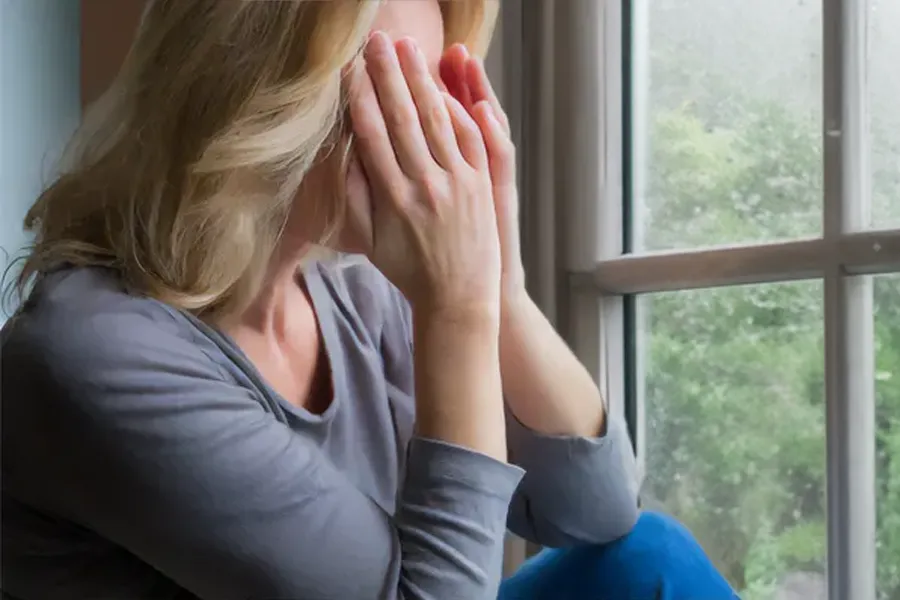 Graves-Disease-Clinic A blonde woman in a gray shirt sits in front of a window on a rainy day covering her eyes with her hands. Get treatment for Grave's Disease from AZENA Health & Longevity in Gainesville.