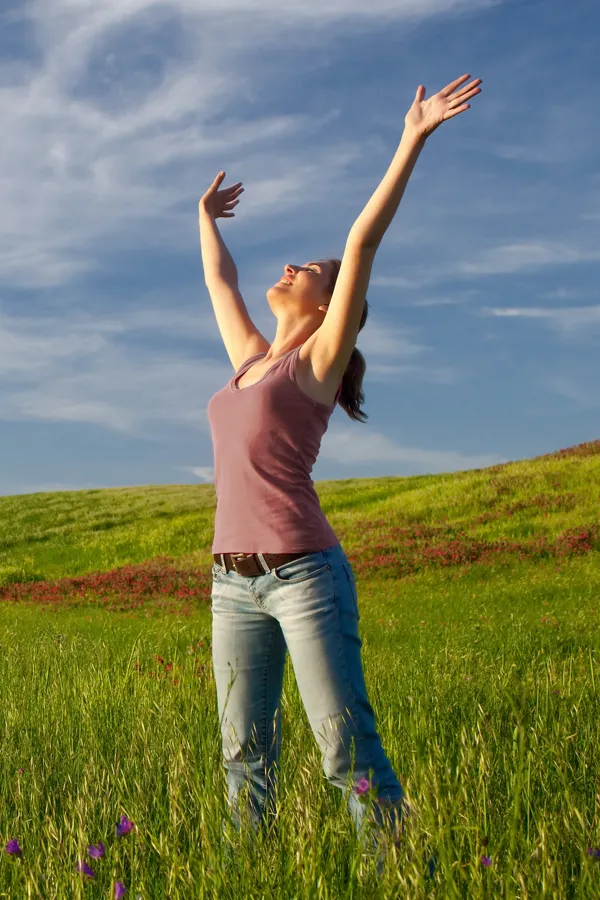 Graves-Disease-Treatment A woman in a tank top and jeans looks up with her arms outstretched to the sky in a field after successful treatment for Grave's Disease from AZENA Health & Longevity in Gainesville.