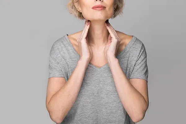 Hashimotos-Thyroiditis-Doctor An older blonde woman in a gray t-shirt holds her hands to her neck checking for signs of thyroid problems. Get help for Hashimoto's Thyroiditis from AZENA Health & Longevity in Gainesville.