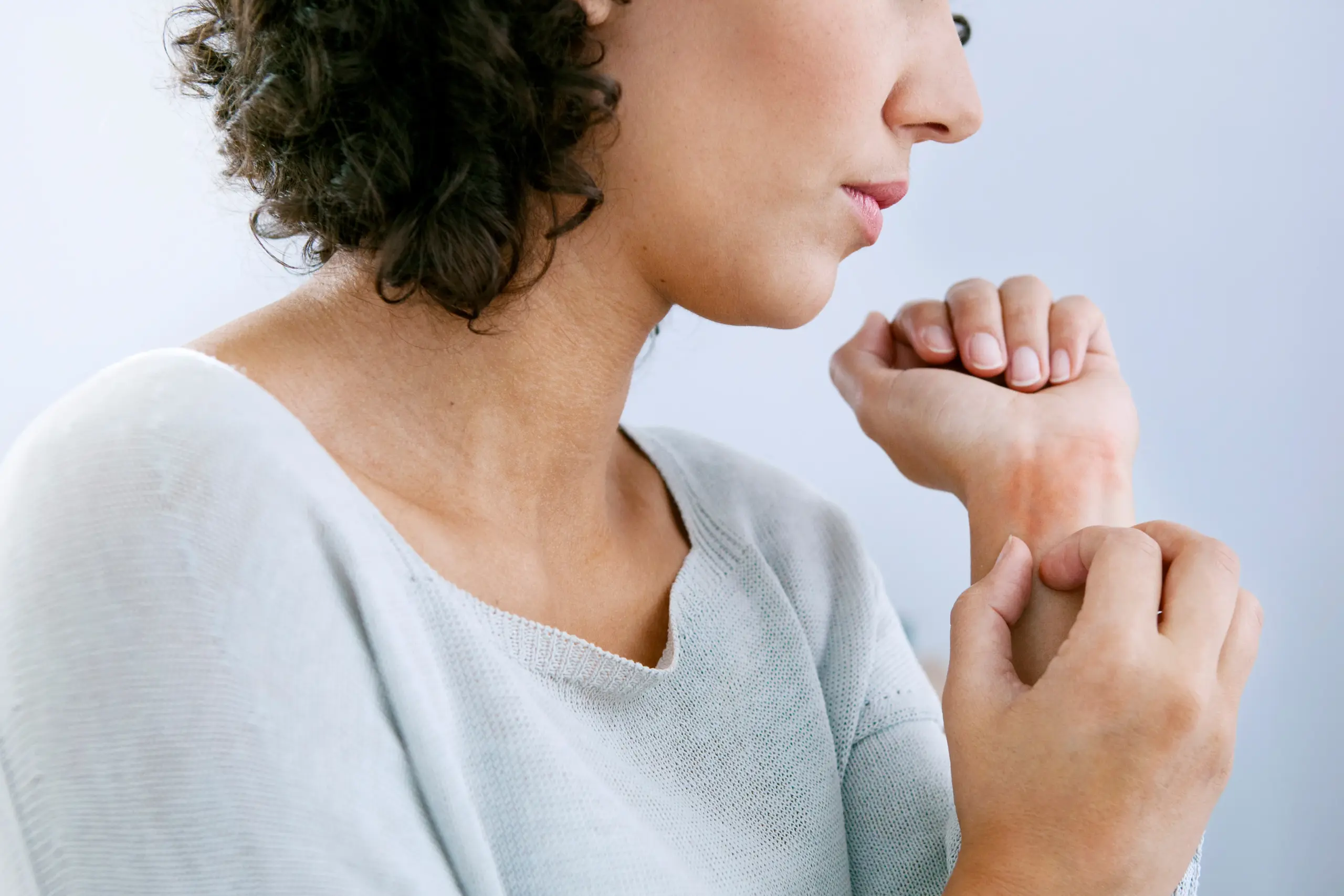 Hives-Doctor A woman with curly brown hair and a light gray shirt scratches her wrist due to hives. Get treatment for hives from AZENA Health & Longevity in Gainesville.