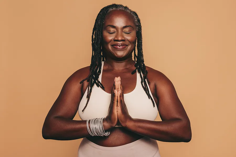 Obesity-Clinic An older African American woman confidently posing against a warm background smiling with hands in a prayer pose. AZENA Health & Longevity treats obesity using functional medicine in Gainesville.