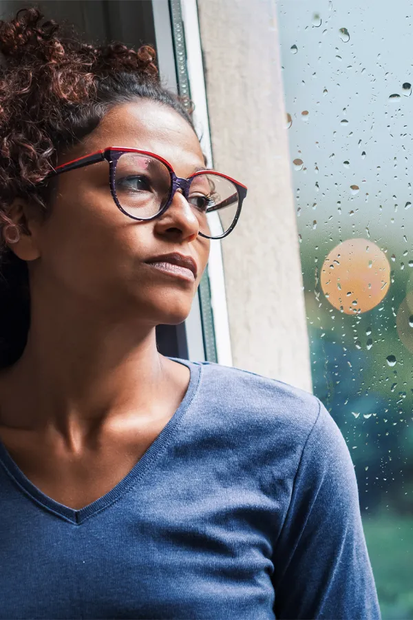 PMDD-Treatment A black woman with with a somber expression and glasses and a gray shirt stares out the window on a rainy day. Get treatment for Premenstrual Dysphoric Disorder from AZENA Health & Longevity in Gainesville.