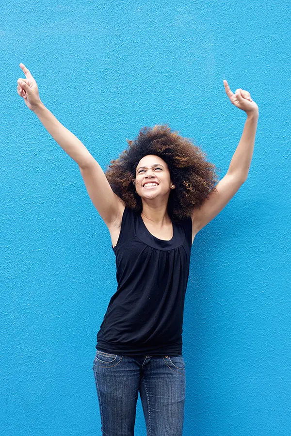 PMS-Treatment A woman in a dark blue tank top standing in front of a bright blue wall, raising her arms in celebration of relief from PMS from AZENA Health & Longevity in Gainesville.