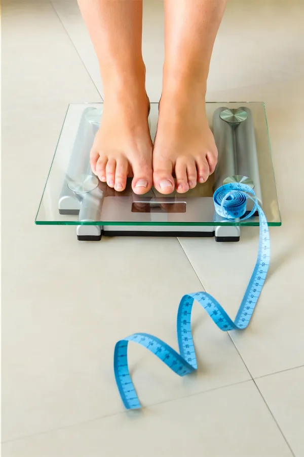 Weight-Loss-Resistance-Treatment Close-up of a woman's feet standing on a scale, with measuring tape by her toes, getting treatment for weight loss resistance from AZENA Health & Longevity in Gainesville.